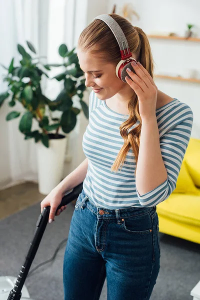 Mulher sorridente ouvir música em fones de ouvido enquanto a limpeza tapete com aspirador de pó em casa — Fotografia de Stock