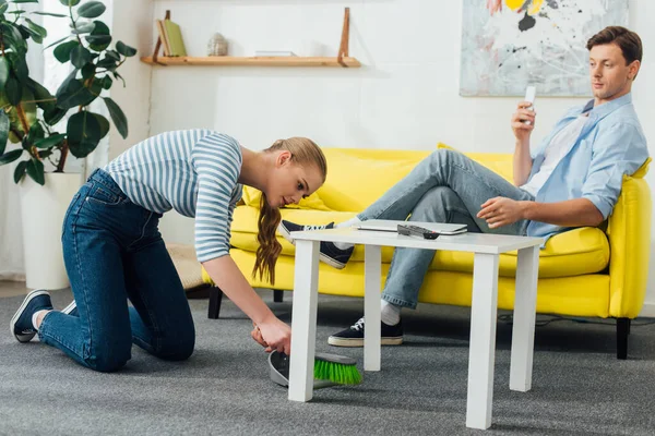 Side view of girl with scoop and broom cleaning carpet near boyfriend using smartphone on couch — Stock Photo