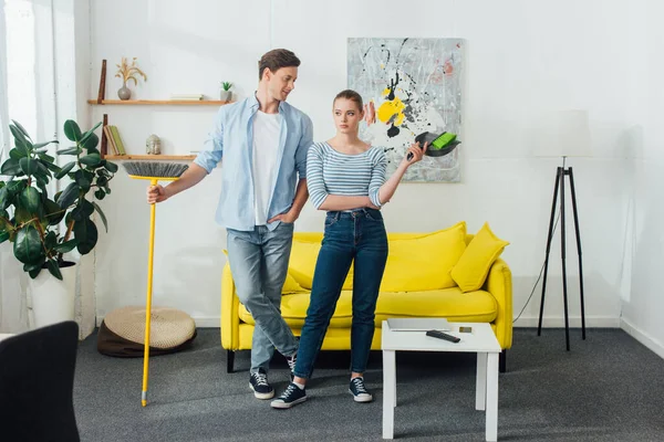 Man looking at girlfriend with scoop and broom in living room — Stock Photo