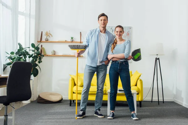Lächelndes Paar mit Schaufel und Besen blickt im Wohnzimmer in die Kamera — Stockfoto