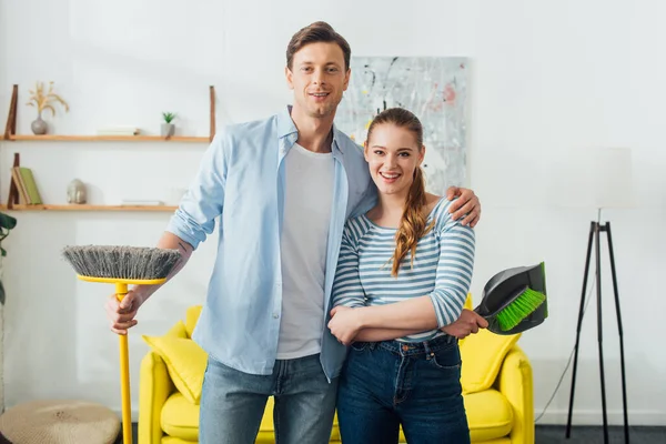 Schöner Mann lächelt in die Kamera, während er Freundin mit Schaufel und Besen im Wohnzimmer umarmt — Stockfoto