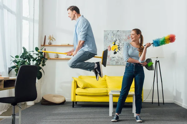 Mujer sonriente con cepillo de polvo y cucharada mirando al novio levitando en la escoba en la sala de estar - foto de stock