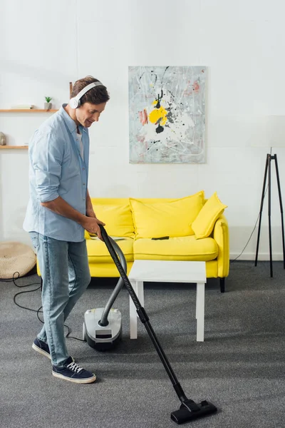 Vista lateral del hombre sonriente en auriculares alfombra de limpieza con aspiradora en casa - foto de stock