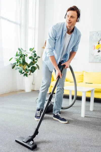 Homem sorridente tapete de limpeza com aspirador de pó e ouvir música em fones de ouvido em casa — Fotografia de Stock