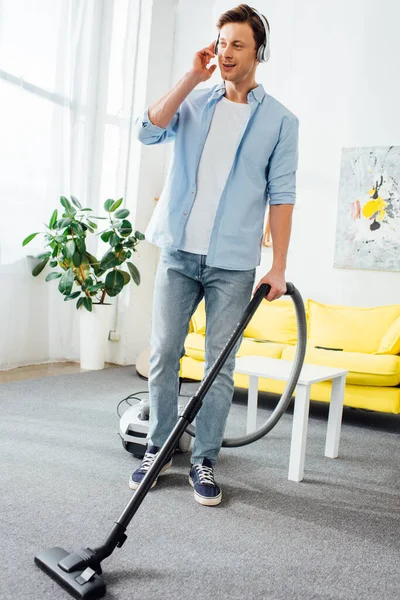 Homme souriant dans les écouteurs nettoyage tapis avec aspirateur dans le salon — Photo de stock