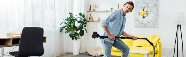 Side view of cheerful man in headphones holding brush of vacuum cleaner in ling room, panoramic shot — Stock Photo