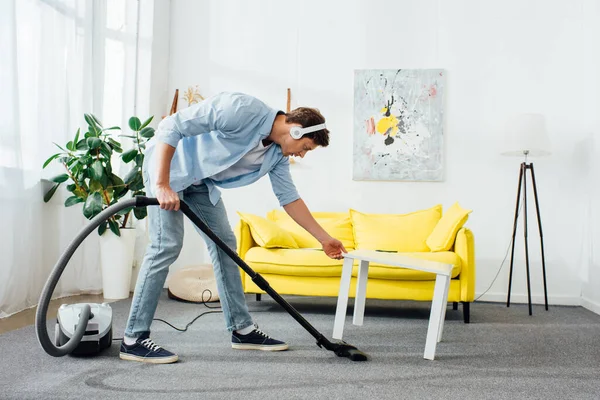 Vista lateral del hombre en auriculares alfombra de limpieza con aspiradora cerca de la mesa de café en casa - foto de stock