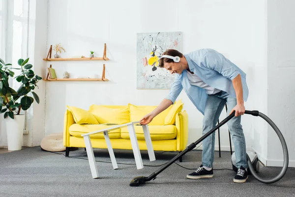 Side view of man in headphones using vacuum cleaner near coffee table at home — Stock Photo