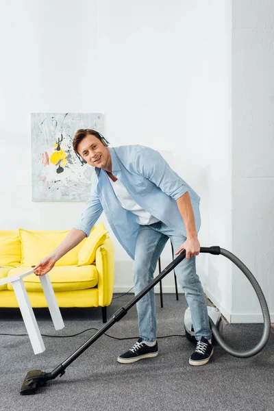 Hombre sonriente en auriculares mirando a la cámara mientras limpia la alfombra con aspiradora en la sala de estar - foto de stock