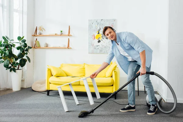 Mann mit Kopfhörer lächelt in die Kamera, während er zu Hause Teppich mit Staubsauger in der Nähe des Couchtisches putzt — Stockfoto