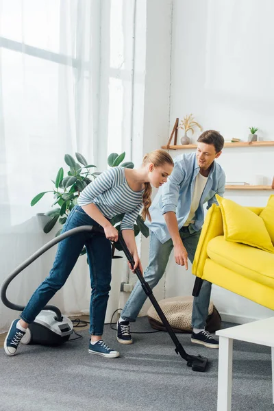Woman cleaning carpet with vacuum cleaner near boyfriend lifting up couch at home — Stock Photo