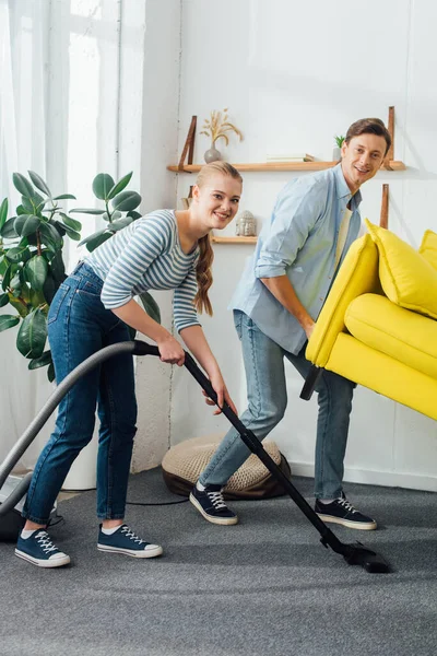 Pareja sonriente mirando a la cámara mientras limpia la alfombra con aspiradora debajo del sofá en la sala de estar - foto de stock