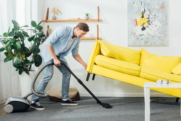 Vue latérale de l'homme soulevant le canapé tout en nettoyant le tapis avec aspirateur dans le salon — Photo de stock