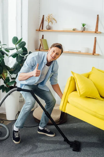 Sorrindo homem mostrando polegar para cima perto aspirador e sofá na sala de estar — Fotografia de Stock