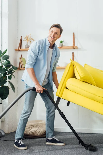 Sorrindo homem olhando para longe enquanto limpando tapete com aspirador de pó sob sofá na sala de estar — Fotografia de Stock