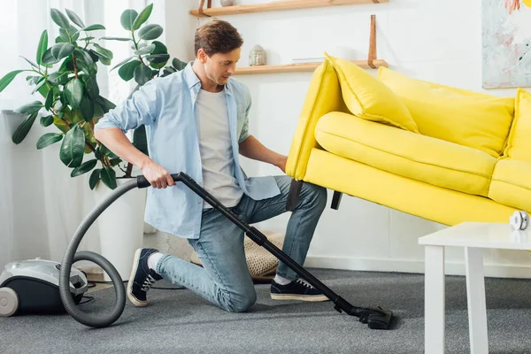 Vue latérale de l'homme soulevant canapé tout en nettoyant le tapis avec aspirateur — Photo de stock