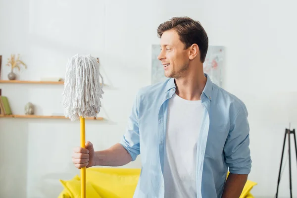 Hombre guapo sonriendo mientras sostiene la fregona en la sala de estar - foto de stock