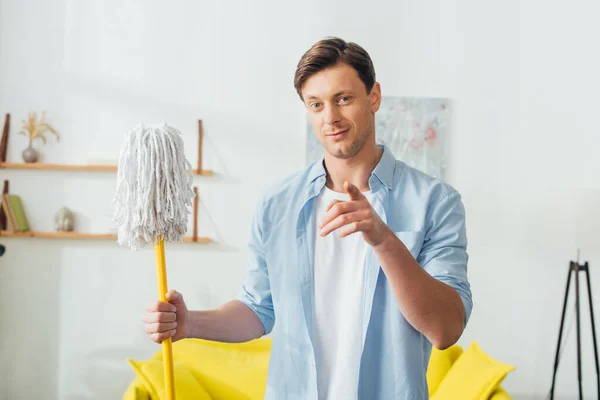 Handsome man pointing at camera and holding mop in living room — Stock Photo