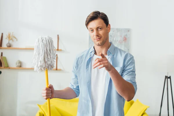 Schöner Mann zeigt auf Kamera und hält Wischmopp im Wohnzimmer — Stockfoto