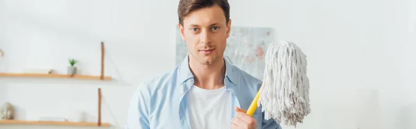 Beau homme regardant la caméra et tenant la serpillière dans le salon, vue panoramique — Photo de stock