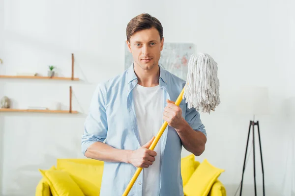 Schöner Mann schaut Kamera an und hält Wischmopp im Wohnzimmer — Stockfoto