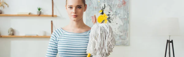 Beautiful woman holding mop and looking at camera in living room, panoramic shot — Stock Photo