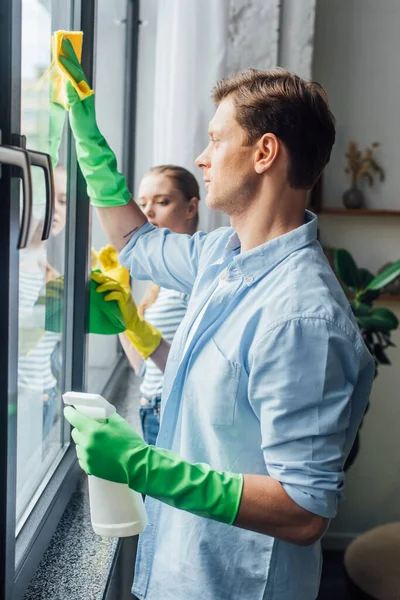 Vue latérale du jeune couple nettoyage verre de fenêtre à la maison — Photo de stock