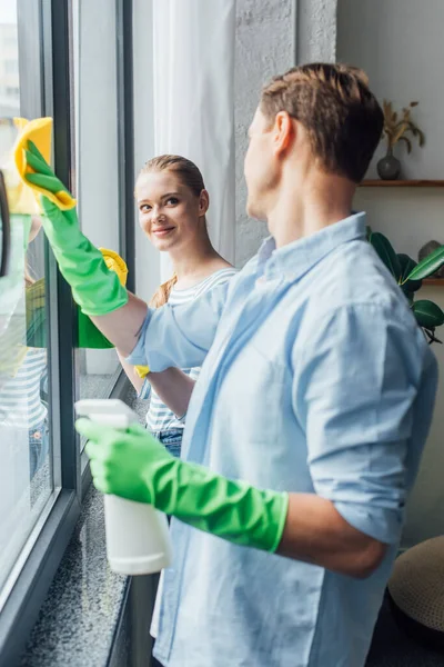 Vista lateral do casal sorrindo um para o outro ao limpar o vidro da janela na sala de estar — Fotografia de Stock