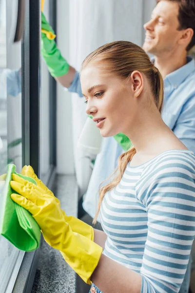 Vue latérale du jeune couple avec chiffons nettoyage fenêtre à la maison — Photo de stock