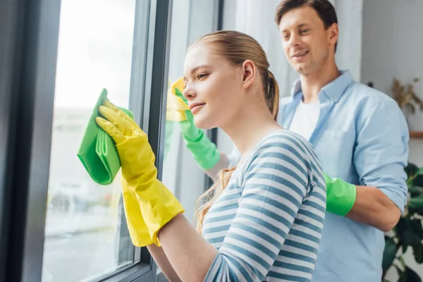 Selective focus of young woman cleaning window near smiling boyfriend in living room — Stock Photo