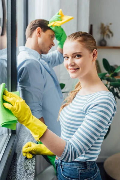 Selektiver Fokus eines Mädchens, das in die Kamera lächelt, während es Fenster putzt, in der Nähe eines müden Freundes mit Lappen im Wohnzimmer — Stockfoto