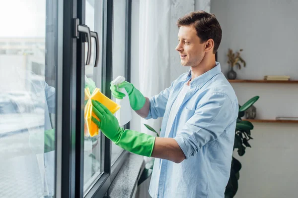 Vista lateral del hombre sonriente con botella de detergente y ventana de limpieza de trapo en casa — Stock Photo