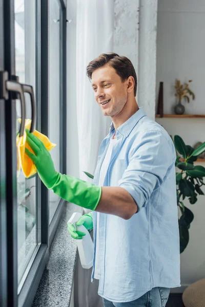 Side view of man with bottle of detergent and rag smiling while cleaning glass of window at home — Stock Photo