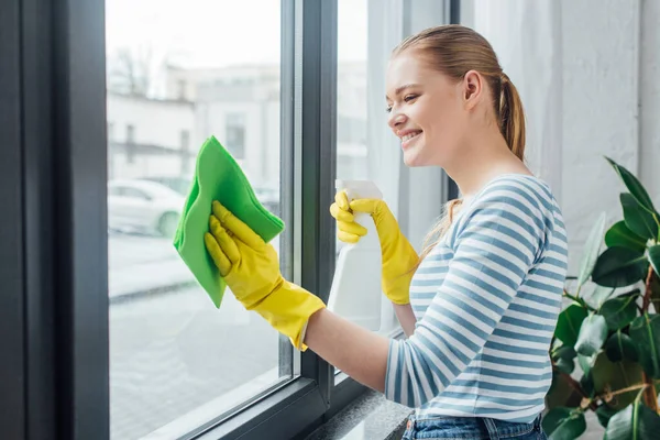Vista lateral de atractiva mujer limpiando vidrio de ventana con trapo y detergente - foto de stock