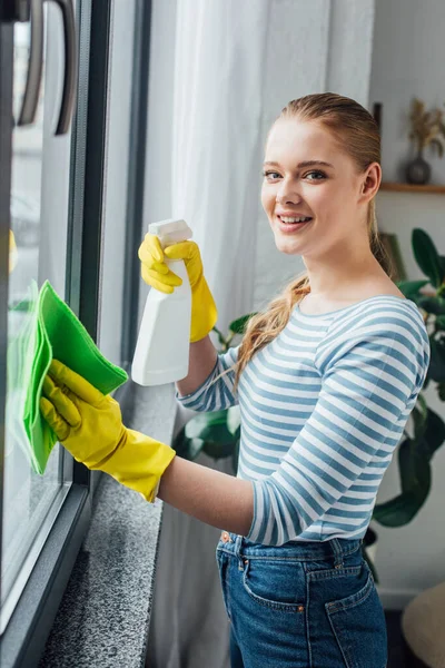 Vista laterale della donna sorridente che guarda la fotocamera mentre pulisce la finestra a casa — Foto stock