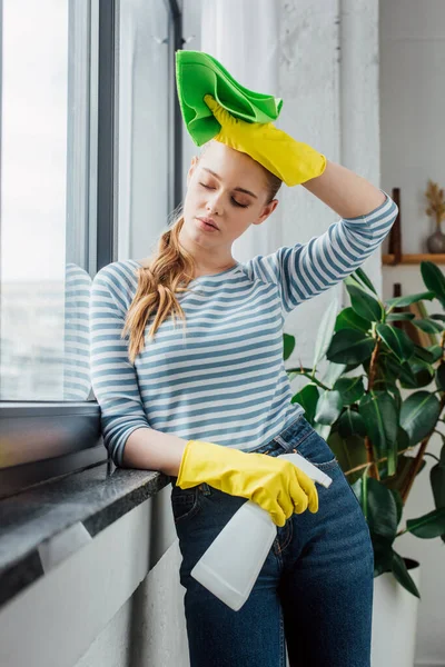 Menina cansada em luvas de borracha segurando detergente e pano perto da janela em casa — Fotografia de Stock