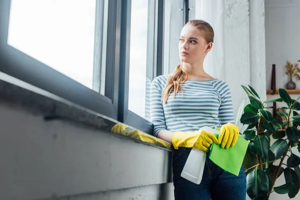 Selective focus of young woman looking away while holding bottle of detergent and rag near window — Stock Photo