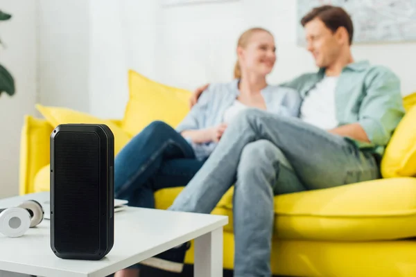 Enfoque selectivo de altavoces inalámbricos y auriculares en la mesa de café cerca de pareja joven en el sofá en casa - foto de stock