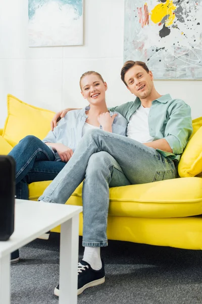 Couple souriant regardant la caméra près de haut-parleur sans fil sur la table basse dans le salon — Photo de stock