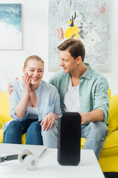 Вибірковий фокус чоловіка, який обіймає усміхнену дівчину на дивані біля бездротового динаміка та навушники на кавовому столі — стокове фото