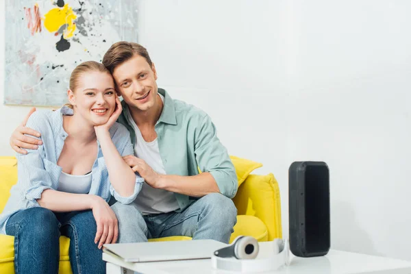 Foyer sélectif de jeune couple souriant à la caméra près de haut-parleur sans fil, écouteurs et ordinateur portable sur la table basse — Photo de stock