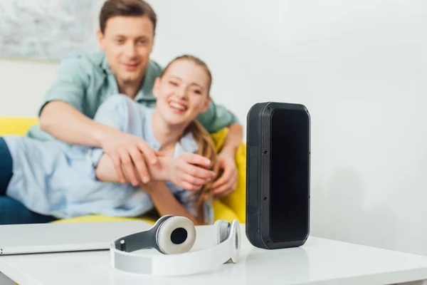 Selective focus of wireless speaker, headphones and laptop on coffee table near cheerful couple on couch at home — Stock Photo