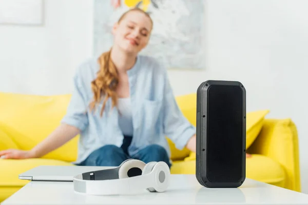 Selective focus of wireless speaker, headphones and laptop on coffee table near smiling woman on couch — Stock Photo