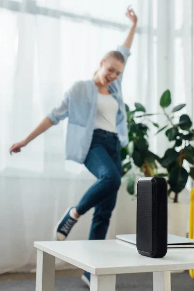 Вибірковий фокус портативного динаміка і ноутбук на кавовому столі і весела жінка танцює у вітальні — стокове фото