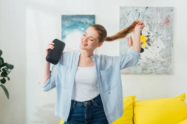 Весела жінка торкається волосся, тримаючи портативний динамік у вітальні — стокове фото