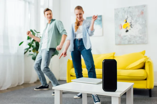 Selektiver Fokus von drahtlosem Lautsprecher und Laptop auf Couchtisch und fröhlichem Paar, das zu Hause tanzt — Stockfoto