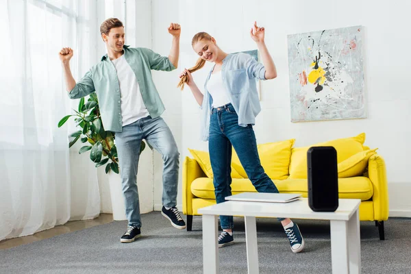 Pareja sonriente bailando cerca de altavoz inalámbrico y portátil en la mesa de centro en la sala de estar - foto de stock