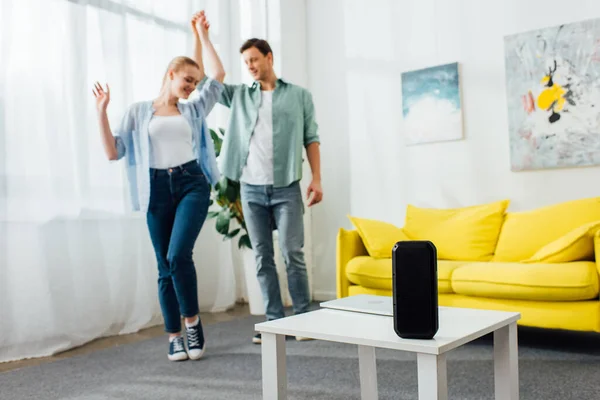 Selective focus of wireless speaker and laptop on coffee table and smiling man dancing with girlfriend at home — Stock Photo