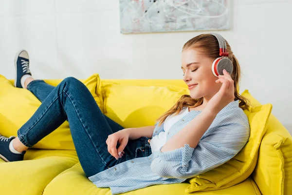 Vista lateral de la chica sonriente en auriculares escuchando música en el sofá - foto de stock