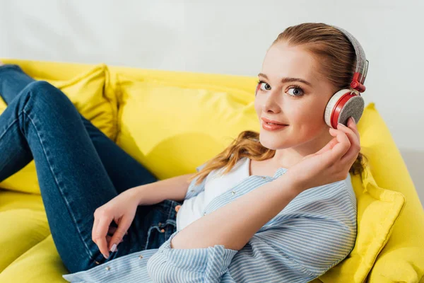 Seitenansicht eines lächelnden Mädchens mit Kopfhörern, das auf der Couch in die Kamera schaut — Stockfoto